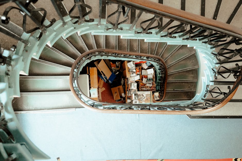 A view looking down a spiraling staircase with a wooden handrail and metal balusters, capturing the staircase as it curves downward into a landing area filled with various packed moving boxes, cardboard cartons, and household items. The boxes are stacked and scattered on the floor, some open revealing contents, while others are sealed with packing tape. There are additional moving supplies such as wrapped furniture or fragile items nearby, indicating a home relocation or furniture transport scenario. The staircase is situated inside a property, with visible steps made of a dark material, possibly wood or laminate, and the landing below is carpeted or mat-covered. The lighting in the image is natural or ambient, highlighting the confined space and the preparations involved in packing and loading during a professional removal process, as conducted by Man with Van Clerkenwell.