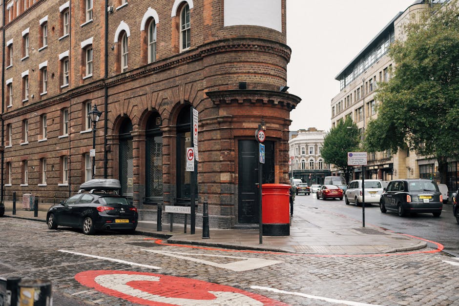 A street scene in Clerkenwell showing a curved red brick building on the corner with arched doorways, black metal bollards, and a red post box near the sidewalk. Several parked cars are visible along the street, including a black car with a roof cargo box parked close to the building. The cobblestone pavement features red and white markings indicating a loading zone. In the background, more parked vehicles and modern office buildings with large windows can be seen, along with some green trees providing partial shade. The scene is well-lit by natural daylight, and the overall environment suggests an urban area suitable for house removals and furniture transport activities by companies like Man with Van Clerkenwell, as part of packing and moving processes. The street appears quiet and organized, with clear signage for parking and loading restrictions.