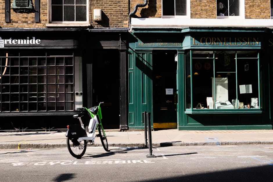 A busy street scene in Clerkenwell showing people walking on the sidewalk beneath a large leafy tree with green foliage. In the foreground, there is a classic red telephone box with the sign 'TELEPHONE' above its glass-paneled door. To the left, a row of bicycles is parked along a bike rack. Behind the pedestrians, there are buildings with brick facades, featuring storefronts and residential balconies, including a sign for a café or bar and a coffee shop. The street is filled with pedestrians dressed in casual clothing, some waiting at a bus stop or crossing the sidewalk. Equipment such as moving blankets, boxes, and wrapping materials is not visible in this scene, indicating it is an external urban environment rather than a home interior. This image depicts an active urban area suitable for considering house removals or furniture transport services, with natural light filtering through the tree's branches. Man with Van Clerkenwell specializes in house removals and relocation services, supporting efficient packing and transport logistics in this vibrant setting.