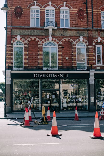 The image shows the facade of a multi-storey red brick building with white decorative arches and window frames, located in an urban area. On the ground floor, there is a shop named 'DIVERTIMENTI' with large glass windows displaying various items, and a black awning with white lettering. In front of the shop, on the sidewalk, multiple orange and white traffic cones are positioned to cordon off a section of the street, possibly indicating ongoing maintenance or a loading zone. A small yellow lifting device with black straps is set on the pavement near the cones, suggesting preparation or ongoing activity related to moving or transport. The scene is illuminated by natural daylight, and the adjacent street appears quiet, with no visible pedestrians or vehicles in motion. This setting reflects typical urban infrastructure involved in home relocation or furniture transport activities, possibly coordinated by a professional removals service like Man with Van Clerkenwell during a home or business move.