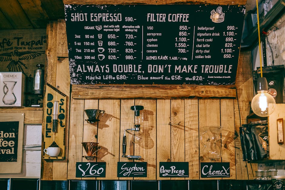 Inside a coffee shop with wooden wall paneling, a black chalkboard menu listing various coffee drinks and prices hangs prominently at the top. Below the menu, there is a coffee brewing station with three pour-over coffee drippers positioned over matching coffee cups, each labeled with handwritten black tags reading '$60', 'Syphon', and 'Aero Press'. To the right, there are glass storage containers and a visible light bulb fixture illuminating the area. The surrounding environment features stainless steel equipment, a framed poster, and additional display boards, with the overall setting suggesting a cozy, rustic space dedicated to coffee preparation and consumption, related to the concept of efficient packing and transport in house removals as part of professional moving services such as those offered by Man with Van Clerkenwell.