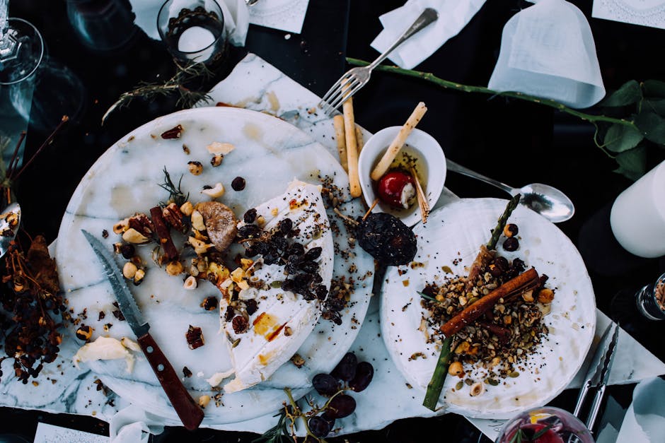 A white marble table holds two partially eaten cheesecakes topped with a variety of nuts, dried fruits, and chocolate bits, with a serrated knife and a dessert fork resting on the desserts. Nearby, there is a small white cup containing a cherry and a wooden stick, along with some scattered grapes and remnants of toppings. The table is surrounded by white napkins, cutlery, and drinking glasses, with a black cloth underneath. The scene appears to be part of a casual dining or celebration setting, with a focus on dessert presentation. This image relates to home removal services by illustrating the importance of careful packing, handling, and transporting delicate items like food, emphasizing the need for professional moving companies such as Man with Van Clerkenwell in the context of house removals and packing and moving logistical support.