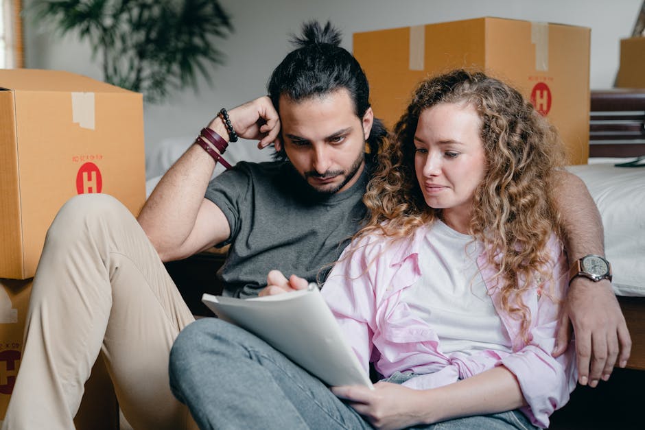 A man with dark hair tied in a top knot, wearing a dark gray T-shirt and beige trousers, sits on the floor next to a woman with curly, light brown hair, wearing a pink shirt. They are surrounded by mo