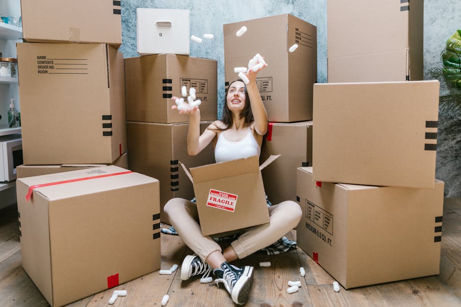 A person wearing light-colored clothing and white socks is inside a room, lifting and placing a cardboard box onto a stack of similar boxes. The boxes are made of brown corrugated cardboard, some of w