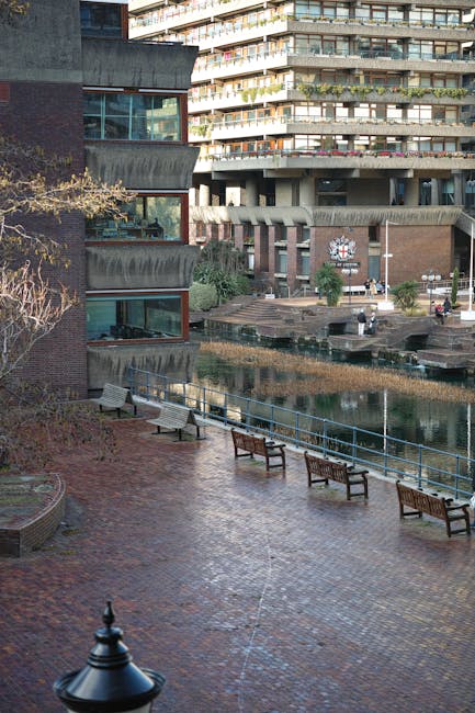The image depicts an outdoor urban area adjacent to a modern residential building with multiple balconies, situated near St John's Square in Clerkenwell. In the foreground, there is a paved pedestrian area with several wooden and metal benches spaced along a curved railing that runs parallel to a small water feature or canal. The paving appears to be brick or stone, with a lamppost visible at the bottom left corner. The residential building has large glass windows and distinctive architectural features, including protruding sections and angled balcony edges. The overall scene is well-lit, with natural daylight illuminating the space. Man with Van Clerkenwell specializes in house removals and furniture transport, and this image reflects the typical moving environment involving loading or unloading processes in an urban setting, where furniture and packing materials might be staged for a home relocation near the Barbican Centre.