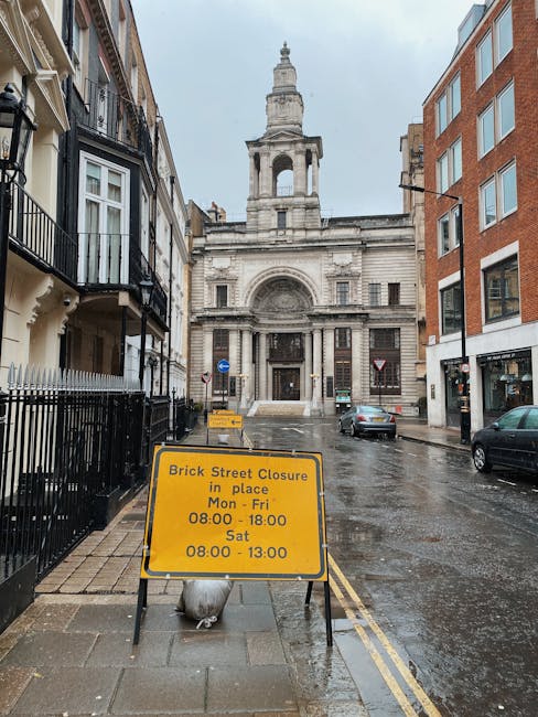 A wet city street in Clerkenwell, London, showing a construction or parking road closure sign placed on the pavement in front of a historic stone building with a large arched entrance, classical architectural details, and a tall tower with a spire. The street is lined with multi-storey residential buildings featuring brick and stucco facades, some with balconies and black wrought-iron railings. Parked cars are visible along the curb, and the road appears to be used for loading or moving activities. The scene suggests ongoing building or home relocation work, with the sign indicating restricted access times for street closures, while the presence of the sign and wet pavement imply recent or ongoing rain, consistent with urban removals and logistics operations typical in house moving services offered by companies like Man with Van Clerkenwell.