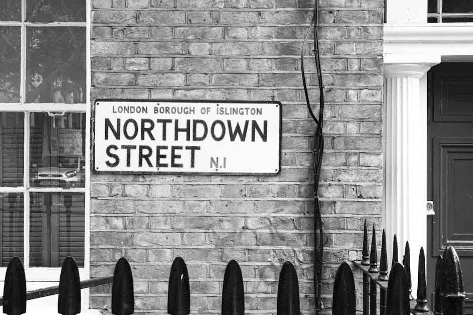 Black and white photograph of a brick building facade on Northdown Street in the London Borough of Islington. A metal street sign is mounted on the wall, displaying 'Northdown Street' with the additional text 'London Borough of Islington' and an abbreviation 'N.I.' Below the sign, a black metal fence with pointed finials is visible at the bottom of the image. To the left of the sign, a portion of a window with a white frame and multiple panes reflects nearby parked cars and a building interior. On the right side, there is a section of a white column supporting an overhang or porch. An exposed black electrical cable runs vertically down the brick wall, slightly to the right of the sign. The lighting appears natural, and the scene is devoid of moving elements, focusing on the building exterior in a typical residential or postcode area, relevant to home relocation and furniture transport logistics. Man with Van Clerkenwell would handle such property possessions during packing or moving processes, often involving loading and unloading in similar environments.