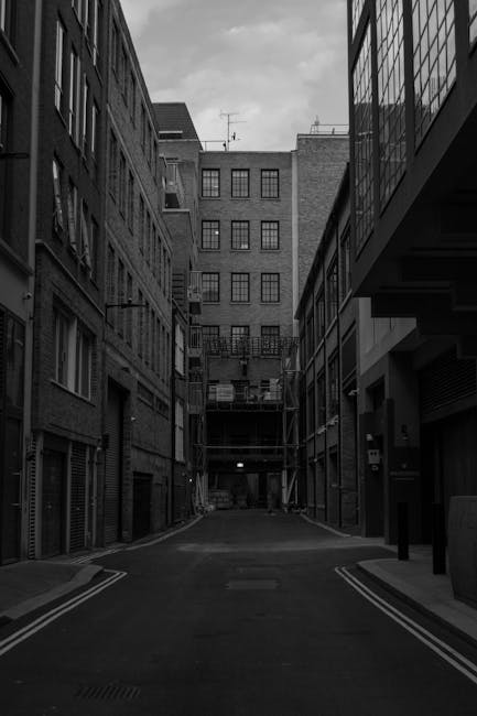 A narrow urban alleyway flanked by multi-storey brick buildings with numerous windows, some with balconies, and a construction scaffold visible at the far end. The alley has a paved surface with double yellow lines along the curb edges, and is illuminated by a few streetlights. The scene suggests a typical city environment suitable for a home relocation or furniture transport process, as managed by Man with Van Clerkenwell, who specialise in removals and moving services in the Clerkenwell area. The image captures the confined space often encountered during moves through narrow streets like Farringdon Road, highlighting the logistical considerations of loading and transporting household furniture and boxes via a commercial van or vehicle suited for urban deliveries.