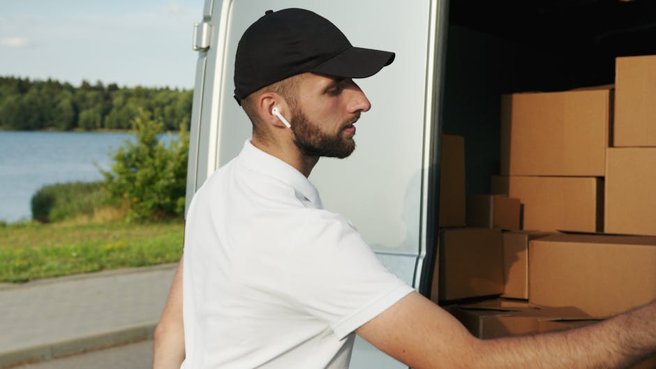 A man wearing a white t-shirt, black cap, and wireless earbuds, engaged in a home relocation task, loading cardboard boxes into the back of a white moving van. The boxes are stacked neatly on the vehi