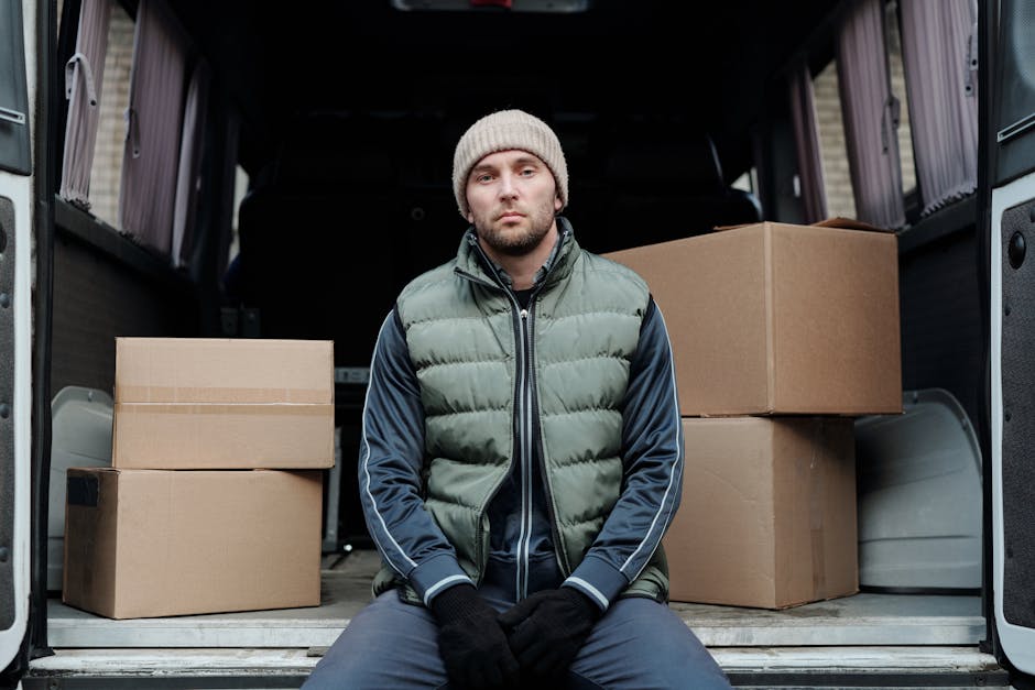 A man wearing a white t-shirt, black cap, and wireless earbuds, engaged in a home relocation task, loading cardboard boxes into the back of a white moving van. The boxes are stacked neatly on the vehi